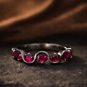 May include: A silver ring featuring seven oval, bright red gemstones set in a flowing, wavy pattern. The ring is presented on a dark, textured surface, with a blurred brown backdrop.