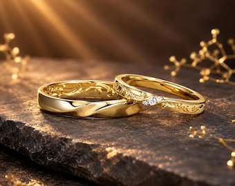 May include: Two gold wedding rings. One ring features a twisted design, while the other has three small diamonds and an engraved floral pattern. The rings are displayed on a dark, textured surface with a warm, golden light.