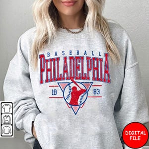 May include: A light grey sweatshirt featuring a distressed red and blue graphic that reads "BASEBALL PHILADELPHIA 18 83". The graphic includes a baseball player silhouette and a baseball. The words "DIGITAL FILE" are in a red circle.