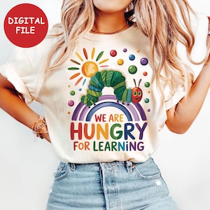 May include: Cream-colored t-shirt featuring a colorful graphic of a caterpillar on a rainbow, with a sun and the text "WE ARE HUNGRY FOR LEARNING." A red circle with "DIGITAL FILE" is in the upper left corner.