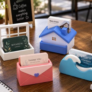 Può includere: Quattro distinti porta biglietti da visita: una cornice bianca, una casa blu con camino, una busta rosa con chiusura a cuore e un'onda blu. Ogni supporto contiene carte con le scritte Botanical Garden, Real Estate, Thank You e Ocean Breeze.