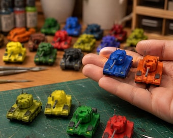 May include: A collection of miniature toy tanks in various solid colours including blue, orange, green, yellow, and red. Each small tank features a rotating turret, a central barrel, and detailed treads. The tanks are displayed on a green mat.