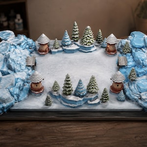 May include: A miniature winter landscape diorama featuring a central snow-covered clearing surrounded by light blue icy rock formations. The scene includes four small wooden towers with conical roofs and several miniature evergreen trees.