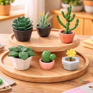 May include: A collection of decorative paper succulents in various pots, arranged on two wooden trays. The succulents are green, with pots in shades of black, white, and orange. The scene includes office supplies like scissors and tape.