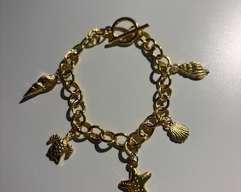 May include: Gold-toned charm bracelet featuring a toggle clasp and various sea-themed charms. The charms include a starfish, seashells, and a turtle. The bracelet is made of linked chain.