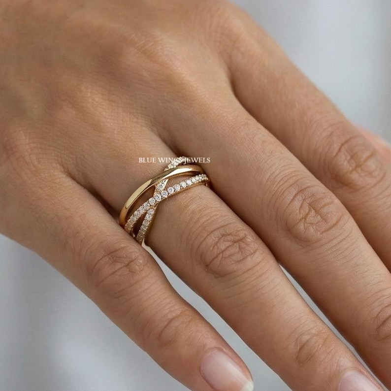 May include: A gold-colored ring with a crisscross design, adorned with small, sparkling stones. The ring features a band that wraps around the finger, creating a layered effect. The ring is on a hand with a light skin tone. The text "BLUE WINGS JEWELS" is visible.