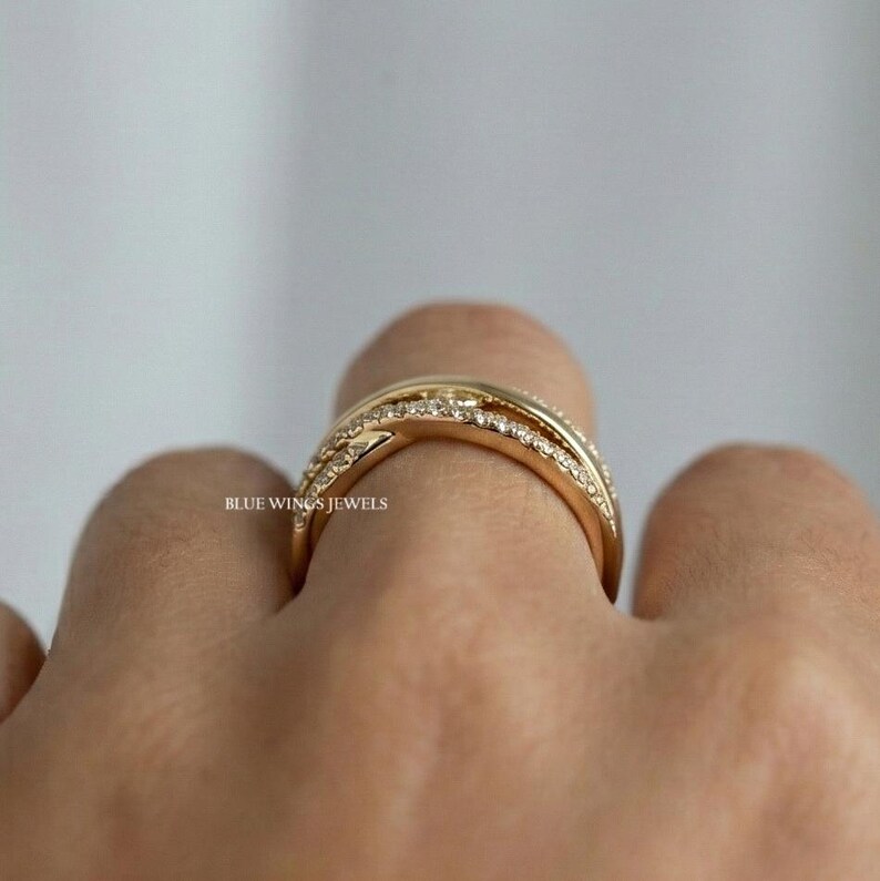 May include: A gold-colored ring with a crisscross design, encrusted with small, sparkling gemstones. The ring is worn on a finger, with the brand name "BLUE WINGS JEWELS" visible.