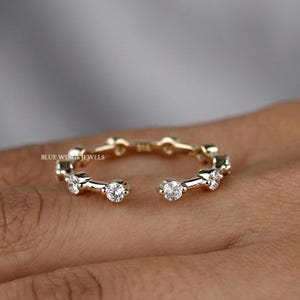 May include: Open-ended gold-toned ring with multiple clear, round gemstones. The ring has a delicate design with a gap in the center. The brand name "BLUE WINGS JEWELS" is visible. This ring is a piece of jewelry.