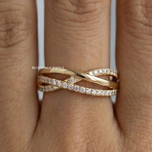 May include: A gold-colored criss-cross ring with small, clear gemstones set along the top band. The ring is on a finger, with the text "BLUE WINGS JEWELS" visible.
