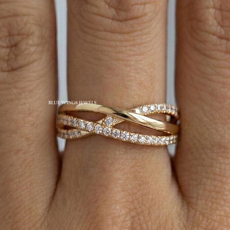 May include: A gold-colored criss-cross ring with small, clear gemstones set along the top band. The ring is on a finger, with the text "BLUE WINGS JEWELS" visible.