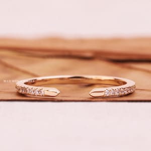 Op de afbeelding: Een goudkleurige open ring met kleine, heldere edelstenen langs de band. De ring heeft een uniek ontwerp met open uiteinden. De tekst "BLUE WINGS JEWELS" is zichtbaar op de ring. De ring wordt gepresenteerd op een houten oppervlak.