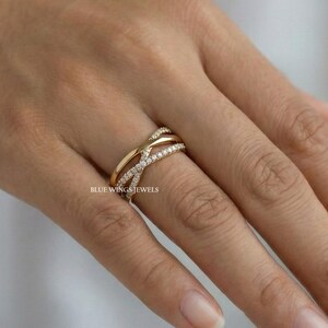 May include: A gold-colored ring with a crisscross design, adorned with small, sparkling gemstones. The ring is on a finger, showcasing its intricate details and elegant style. The text "BLUE WINGS JEWELS" is visible.