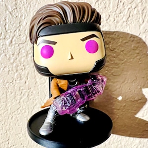Könnte beinhalten: Eine Funko Pop! Vinylfigur der Marvel Comics-Figur Gambit. Die Figur hat ein hellbraunes Gesicht, braunes Haar und ein schwarzes Stirnband. Er trägt eine braune Jacke, silberne Stiefel und hält einen durchscheinenden lila Energieeffekt. Die Figur steht auf einem schwarzen, kreisförmigen Sockel.