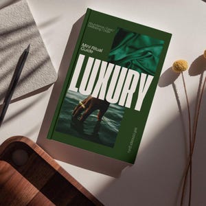 May include: A green book titled "LUXURY" with the text "Mini Ritual Guide" and "Abundance / Ease / Wellbeing / Order." The cover features a hand with rings in water and green fabric. A pencil and wooden tray are also visible.