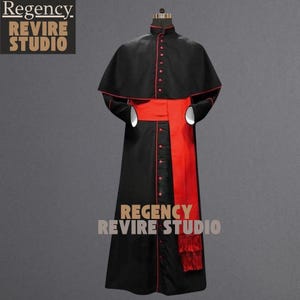 May include: A black and red clerical robe with a matching cape. The robe features red buttons and trim, a red sash, and a fringed detail. The Regency Revire Studio logo is visible.