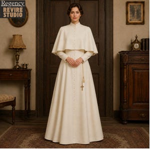Könnte beinhalten: Ein cremefarbenes, geknöpftes Kleid mit passendem Cape. Das Kleid hat lange Ärmel und einen hohen Kragen. Ein goldfarbener Kreuzanhänger hängt an einer goldfarbenen Kette. Das Bild stammt von Regency Revire Studio.