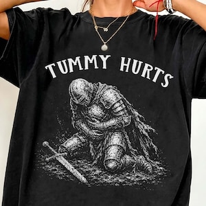 Może przedstawiać: Czarny t-shirt z krótkim rękawem z białym nadrukiem graficznym przedstawiającym rycerza w pełnej zbroi klęczącego na ziemi z mieczem. Napis "TUMMY HURTS" jest nadrukowany białymi wielkimi literami w łuk nad ilustracją rycerza.