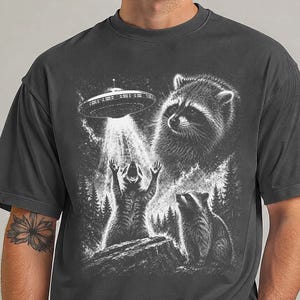 Könnte beinhalten: Ein anthrazitfarbenes Kurzarm-T-Shirt mit einem weißen Grafikdruck von zwei Waschbären in einem Wald. Ein Waschbär steht mit erhobenen Pfoten unter einem schwebenden UFO, das einen Lichtstrahl aussendet, während ein zweiter Waschbär daneben steht.