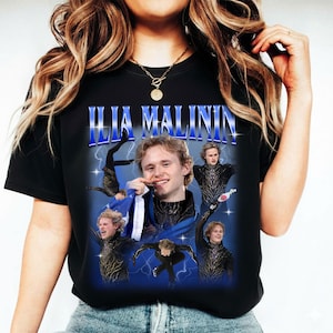 May include: Black t-shirt featuring a graphic of Ilia Malinin with the text "ILIA MALININ" in blue. The design includes multiple images of the skater in action, set against a blue background with lightning bolts.