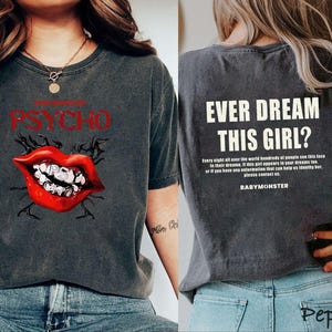 May include: Dark gray t-shirt with the text "PSYCHO" above a red lip graphic with white teeth. The back of the shirt reads "EVER DREAM THIS GIRL?" with additional text and the name "BABYMONSTER".