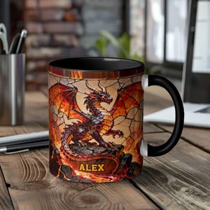 Può includere: Tazza da caffè con manico e interno neri, decorata con un disegno di drago rosso e arancione. Il drago è raffigurato con grandi ali e siede su una superficie rocciosa, con il nome "ALEX". La tazza è su un tavolo di legno.