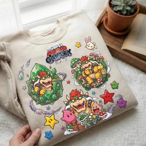 Puede incluir: Sudadera beige con gráficos coloridos de "The Super Mario Galaxy Movie". El diseño incluye personajes de Bowser, estrellas, setas y otros elementos del juego. El texto "The Super Mario Galaxy Movie" también es visible.