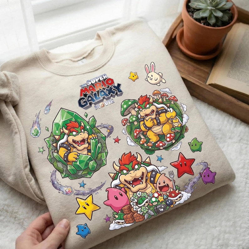 Puede incluir: Sudadera beige con gr&aacute;ficos coloridos de "The Super Mario Galaxy Movie". El dise&ntilde;o incluye personajes de Bowser, estrellas, setas y otros elementos del juego. El texto "The Super Mario Galaxy Movie" tambi&eacute;n es visible.