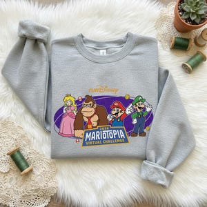 Puede incluir: Una sudadera gris con un gráfico colorido de Mario, Luigi, la Princesa Peach y Donkey Kong. El diseño incluye el texto "runDisney" y "MARIOTOPIA VIRTUAL CHALLENGE 2026". La sudadera se muestra sobre una superficie blanca y esponjosa.
