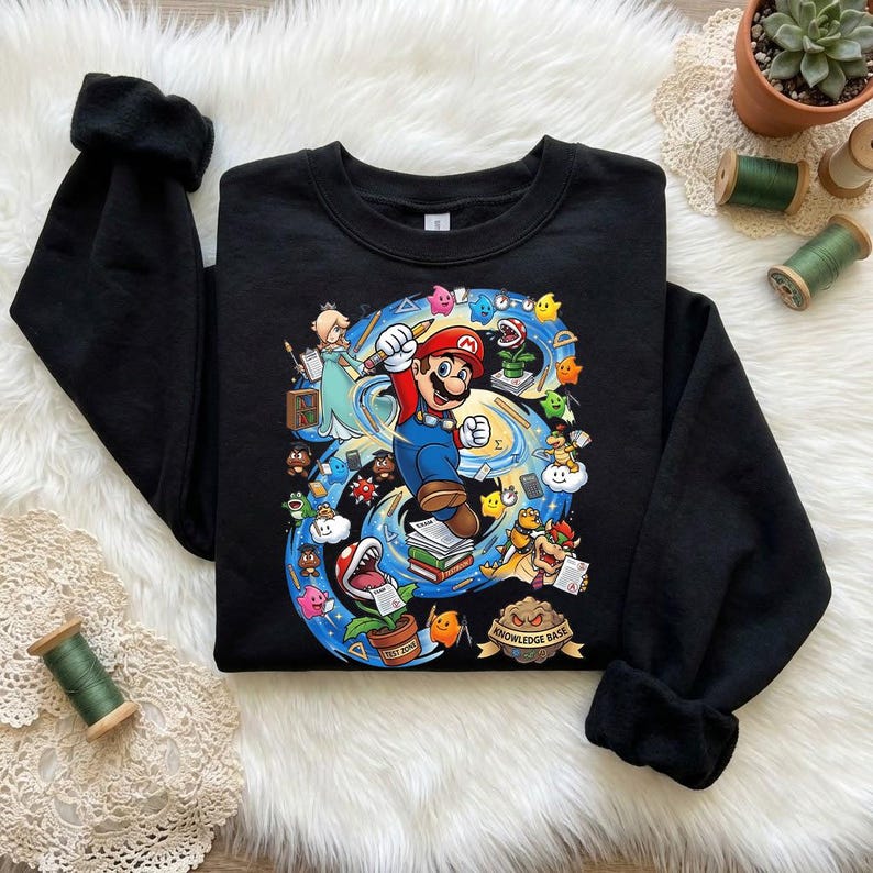 Puede incluir: Sudadera negra con un gr&aacute;fico colorido de Mario del videojuego Super Mario Bros. El dise&ntilde;o muestra a Mario con un l&aacute;piz, rodeado de otros personajes y elementos del juego. Tambi&eacute;n se ve el texto "Knowledge Base".