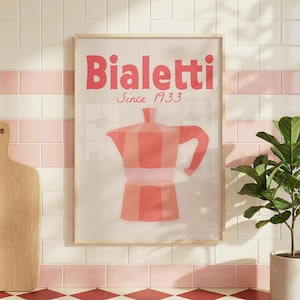 May include: Framed print with a pink Bialetti coffee maker illustration. The text "Bialetti Since 1933" is displayed in pink. The artwork is set against a white background with a light wooden frame, suitable for kitchen decor.
