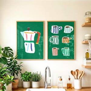 Puede incluir: Dos impresiones artísticas enmarcadas con ilustraciones de temática cafetera. Una impresión muestra una cafetera blanca y colorida sobre un fondo verde. La otra muestra seis tazas de café coloridas con diseños de rayas. Ambas impresiones están en marcos de madera.