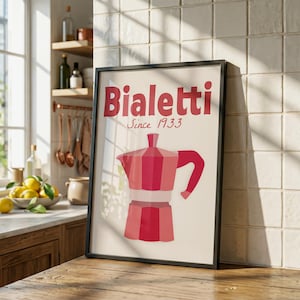 May include: Framed art print featuring a red and pink Bialetti stovetop espresso maker illustration. The text "Bialetti Since 1933" is displayed above the coffee maker image. The print is in a black frame, set on a wooden surface.