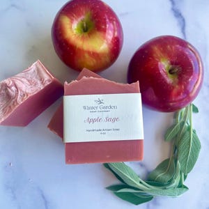 May include: A rectangular bar of pink soap with a white paper label reading Winter Garden Soap Company Apple Sage Handmade Artisan Soap 4 oz, accompanied by two shiny red apples and a sprig of fresh green sage leaves arranged around the soap.
