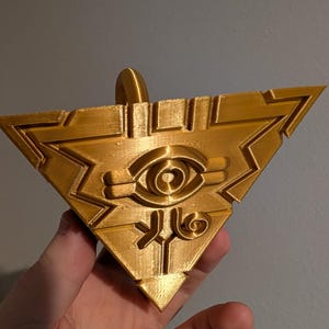May include: A gold-colored, triangular pendant with a raised eye design in the center. The pendant has a loop at the top for hanging and features intricate geometric patterns. The overall design is reminiscent of a stylized emblem or symbol.
