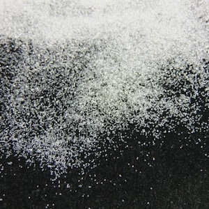 May include: Close-up of a scattering of small, clear, crystalline particles against a dark background. The particles are densely concentrated in the upper portion of the image, gradually thinning out towards the bottom.