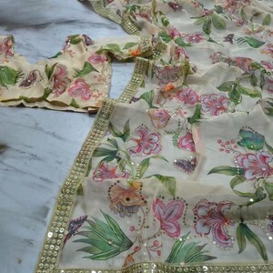 May include: A cream-colored floral saree with a matching blouse. The saree and blouse feature a vibrant print of pink and orange flowers, green leaves, and butterflies. The edges are trimmed with gold sequins.