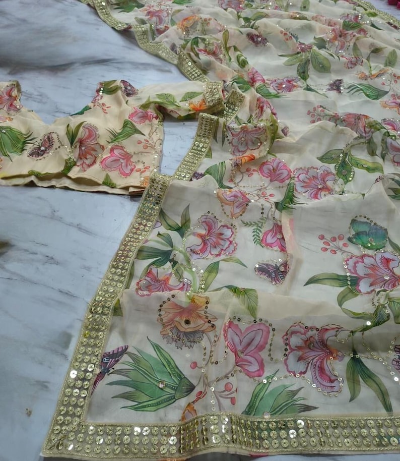 May include: A cream-colored floral saree with a matching blouse. The saree and blouse feature a vibrant print of pink and orange flowers, green leaves, and butterflies. The edges are trimmed with gold sequins.