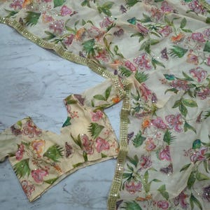 May include: A cream-colored floral saree and matching blouse set. The saree and blouse feature a delicate pattern of pink and peach flowers with green leaves and butterflies. The saree has a gold sequin border.