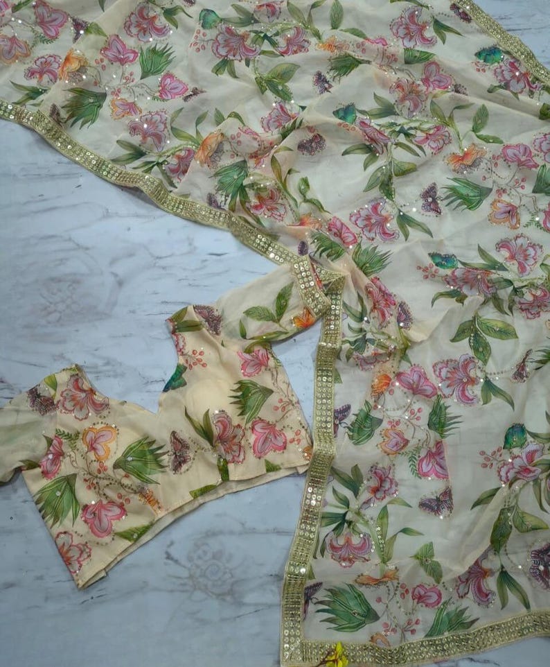 May include: A cream-colored floral saree and matching blouse set. The saree and blouse feature a delicate pattern of pink and peach flowers with green leaves and butterflies. The saree has a gold sequin border.