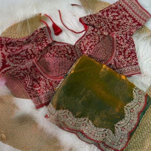 May include: A red embroidered blouse with a deep V-neck and matching sleeves. The blouse features intricate floral patterns and is paired with a green and gold sheer fabric with a decorative border. The items are displayed on a white fluffy surface.