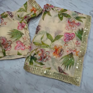 May include: A cream-colored saree and blouse set with a floral pattern in pink, orange, and green. The saree has a gold sequined border. The blouse has short sleeves and a matching floral design.