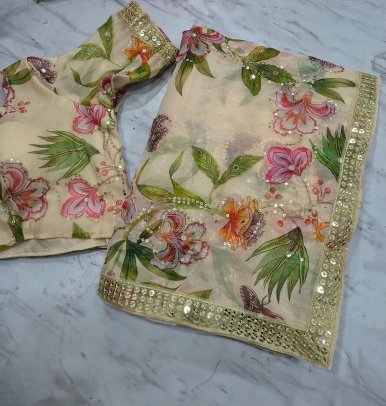 May include: A cream-colored saree and blouse set with a floral pattern in pink, orange, and green. The saree has a gold sequined border. The blouse has short sleeves and a matching floral design.
