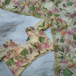 May include: A cream-colored floral saree and blouse set. The saree and blouse feature a delicate pattern of pink, orange, and green flowers with sequin accents. The saree has a gold border. The blouse has short sleeves.
