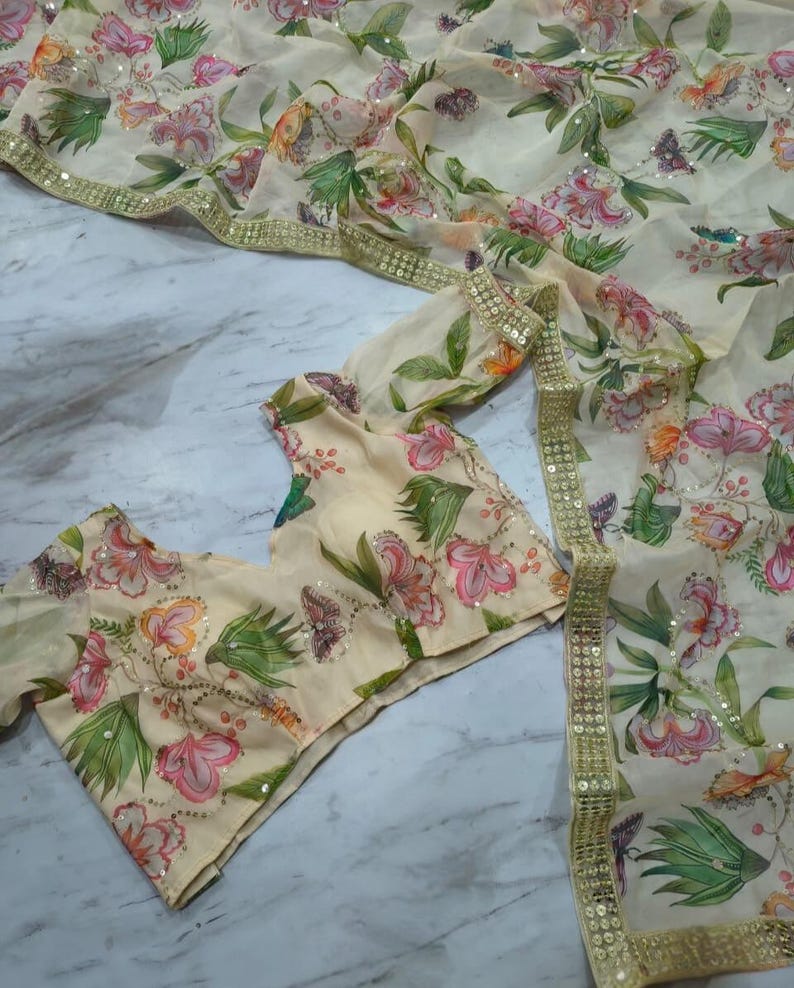 May include: A cream-colored floral saree and blouse set. The saree and blouse feature a delicate pattern of pink, orange, and green flowers with sequin accents. The saree has a gold border. The blouse has short sleeves.