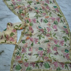 May include: A cream-colored floral saree with a matching blouse. The saree features a delicate pattern of pink and green flowers with gold sequin trim along the edges. The fabric is lightweight and flowing.