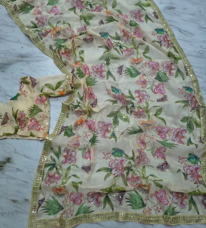 May include: A cream-colored floral saree with a matching blouse. The saree features a delicate pattern of pink and green flowers with gold sequin trim along the edges. The fabric is lightweight and flowing.