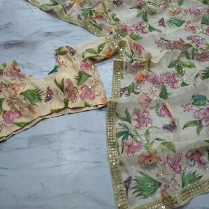 May include: A cream-colored floral saree set with a matching blouse. The saree and blouse feature a delicate pattern of pink and orange flowers, green leaves, and butterflies. The edges are trimmed with gold sequins.