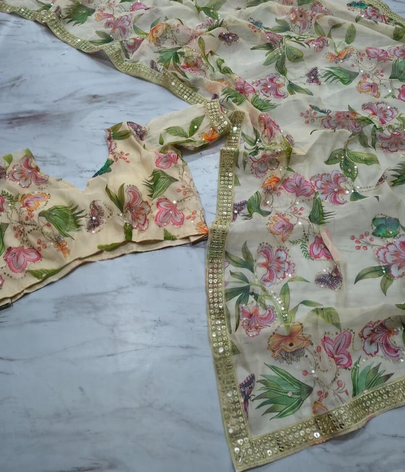 May include: A cream-colored floral saree set with a matching blouse. The saree and blouse feature a delicate pattern of pink and orange flowers, green leaves, and butterflies. The edges are trimmed with gold sequins.