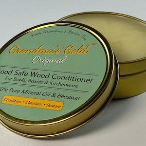 May include: A gold tin with a green label that reads "Grandma's Gold Original Food Safe Wood Conditioner For Bowls, Boards & Kitchenware 100% Pure Mineral Oil & Beeswax Condition Maintain Restore". The tin is open and the inside is filled with a white, waxy substance.