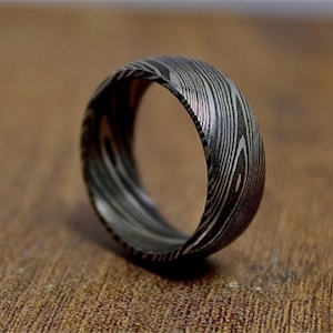 Peut inclure: Gros plan d'une bague en acier de Damas. La bague présente un motif unique de lignes tourbillonnantes dans des tons de gris et de noir, ressemblant à du grain de bois. La bague est posée sur un fond marron flou.